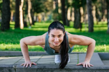 Sportif ve güler yüzlü genç bayan sporcu spor giyim koçu parkta şınav çekiyor. Spor açık hava konsepti.