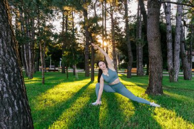 Sportif ve güler yüzlü genç bayan spor giyim koçu yeşil yaz parkında fiziksel esneme egzersizleri yapıyor. Spor açık hava konsepti.