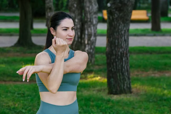 stock image Sportive and smiley athletic young female fitness coach in sportswear doing physical stretch exercises in green summer park. Sport outdoors concept.