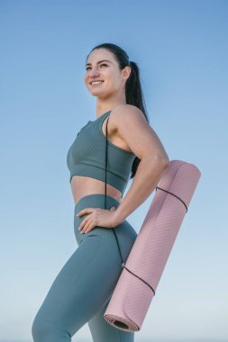 Sportif ve güler yüzlü spor giyim koçu yoga minderini tutuyor ve plajda duruyor. Yoga ve esneme konsepti.