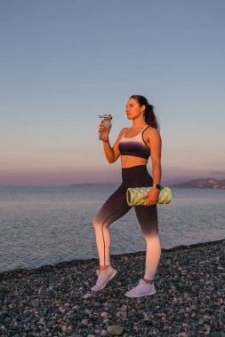 Sportif ve gülen atletik spor koçu spor kıyafetleri giyip su şişesinden su içiyor. Elinde fitness rulosu var. Gün batımında çekilen spor konsepti..