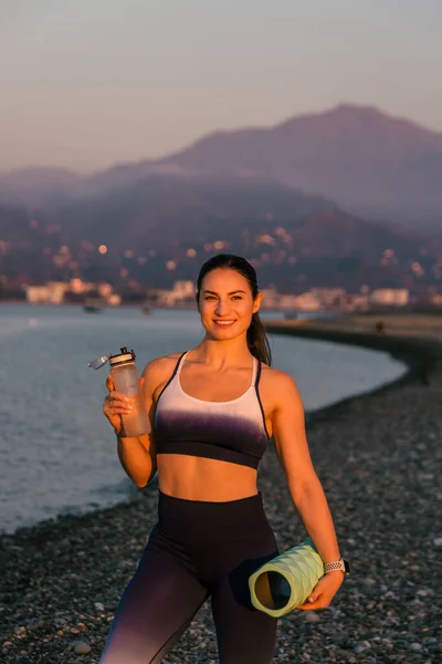 ウォーターボトルからスポーツウェア飲料水を身に着けているスポーティでスマイリー運動若いフィットネスコーチ 彼女は手にフィットネスロールを持っている 美しい夕日の間に撮影されたスポーツ屋外のコンセプト — ストック写真