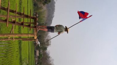 Yeşil pirinç tarlasının üzerindeki tahta köprüde bir adam duruyor. Asya tarzı konik şapka takıyor ve Lao bayrağını taşıyor..