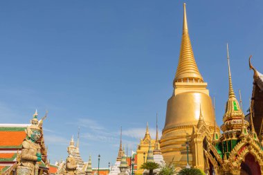 Wat Phra Kaew tapınağının altın kuleleri ve muhafızları, karışık desenlerle bezenmiş, güneşli bir günde mavi gökyüzü, Bangkok, Tayland
