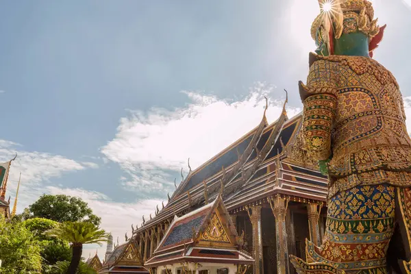 stock image Wat Phra Kaew temple, richly adorned with ornament and gold, against a blue sky on a sunny day, with a guardian statue facing away in the foreground, Bangkok, Thailand