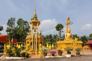 A golden Buddha statue and ornate golden pavilion with bells, surrounded by lush greenery and flowers, at Wat Thong Klang Temple in Thailand clipart