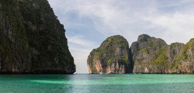 Maya Bay on Phi Phi Ley Island, Thailand, featuring towering limestone cliffs covered in lush greenery surrounding turquoise waters under a clear sky clipart