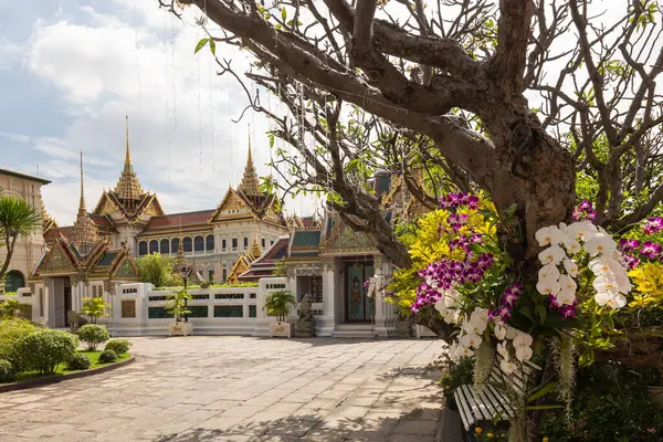 stock image View of the ornately decorated and golden Chakri Maha Prasat, with trees and flowers in the foreground on a sunny day, Bangkok, Thailand