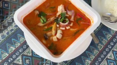 Karides, mantar ve taze otlarla dolu canlı bir kase Tayland Tom Yam çorbası, geleneksel Tayland mutfağının zengin ve baharatlı lezzetlerini gösteren beyaz pirinç tabağının yanında servis edilir..