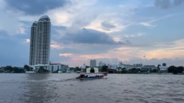 Geleneksel bir nehir teknesi gün batımında Bangkok 'taki Chao Phraya Nehri boyunca yol alır. Yüksek binalar ve renkli bir akşam gökyüzü manzaralı kentsel zemin sağlar..