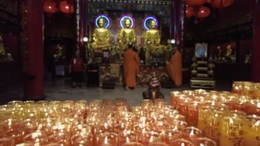 Turuncu cüppeli keşişler, altın Buda heykelleriyle süslenmiş bir tapınakta ayin yapıyorlar. Ön planda yanan mum sıraları parlıyor. Bangkok, Tayland - 25 Ekim 2023.