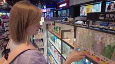 Sırt çantalı genç bir kadın, iyi aydınlatılmış bir güzellik mağazasında cilt ürünlerine göz atıyor. Güzelce düzenlenmiş kozmetiklerle çevrili bir şişeyi dikkatle inceliyor. Bangkok, Tayland - 29 Ekim 2023.