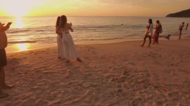 İki kadın gün batımında kumsalda sakin bir anı yakalıyor, kumdan ve sudan yansıyan altın ışıkla, huzurlu ve resimsel bir manzara yaratıyor. Phuket, Tayland - 15 Kasım 2023.