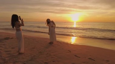 İki kadın gün batımında kumsalda sakin bir anı yakalıyor, kumdan ve sudan yansıyan altın ışıkla, huzurlu ve resimsel bir manzara yaratıyor. Phuket, Tayland - 15 Kasım 2023.