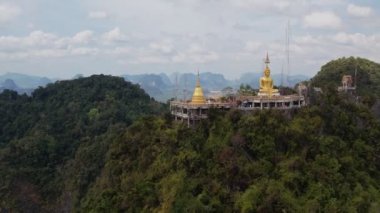 Tayland, Krabi 'deki yemyeşil ormanların ve dramatik kireçtaşı kayalıklarının ortasındaki bir dağın tepesine tünemiş Kaplan Mağarası Tapınağı Wat Tham Suea' daki Altın Buda heykelinin hava manzarası..