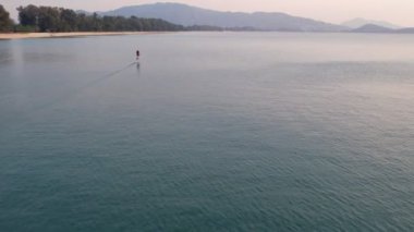 Kürekçi sakin denizde süzülüyor, uzak tepeler ve arka planda sakin bir kıyı şeridi ile, o anın huzurunu yakalıyor. Phuket, Tayland - 28 Ekim 2023.