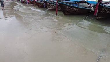 İnsanlar kıyı şeridi boyunca yürürken, arka planda yüksek kireçtaşı kayalıkları olan kumlu bir plajda renkli uzun kuyruklu tekneler sıralanır. Krabi, Tayland - 2 Ocak 2023.