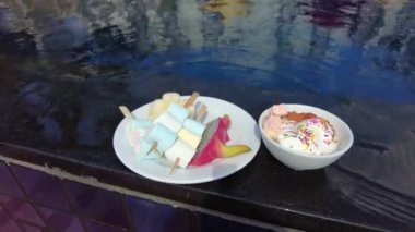 Bir tabak renkli atıştırmalık ve havuz kenarında serpiştirilmiş bir kase dondurma rahatlatıcı ve hoşgörülü bir tatil anını yansıtır..