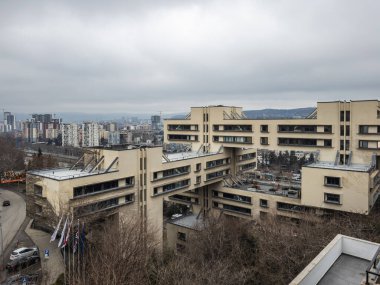 Birbirine bağlı blokları olan modern bir mimari yapı bulutlu bir gökyüzü altında sayısız yüksek binalara sahip bir şehir manzarasına karşı kurulmuş..