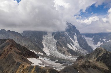 Bulutlu bir atmosferde yükselen zirvelerin arasına yerleştirilmiş bir buzulla engebeli dağlık arazinin çarpıcı bir görüntüsü..