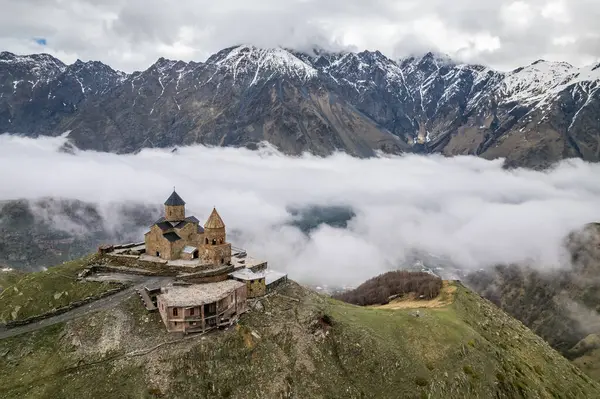 stock image Aerial drone view of Gergeti Trinity Church with heavy fog clouds flowing over the mountain. Beautiful mountain view of the snow-capped peaks near Stepantsminda, Georgia.