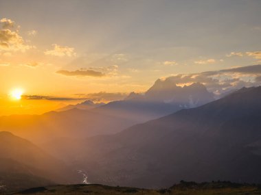 A mesmerizing sunset view over the Caucasus Mountains, casting warm hues and silhouettes across the peaks. Soft clouds scatter the sky, enhancing the golden glow and creating a serene landscape. clipart