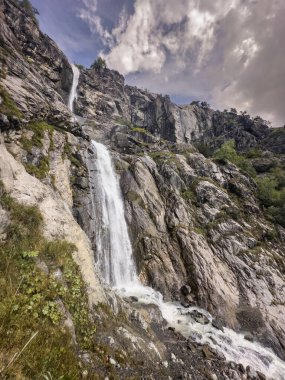 A powerful waterfall descends from a steep, rocky cliff, surrounded by patches of green vegetation and rugged rock formations. clipart