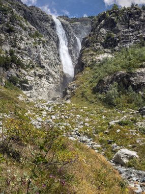 Güçlü bir şelale dikey olarak yeşil bitki örtüleriyle çevrili engebeli kayalık bir yamaçtan aşağı iner.