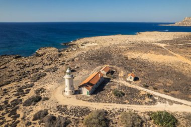 A remote lighthouse surrounded by rugged coastal terrain overlooking the vast, calm sea, captured from above. clipart