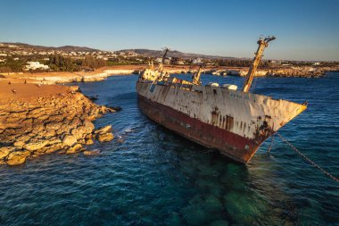 Paslı bir gemi enkazı kayalık bir sahil şeridinde dinleniyor, yıpranmış yapısını bir kıyı kasabası ve sakin sulara karşı sergiliyor. Paphos, Kıbrıs - 27 Ekim 2024.
