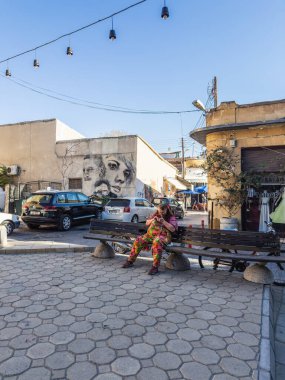 A relaxed street view featuring a woman on a bench, vibrant graffiti artwork, parked cars, and hanging lights add charm to the urban atmosphere. Nicosia, Cyprus - October 26, 2024. clipart