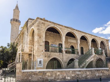 Üzerinde yeşil demir parmaklıklar ve merdivenler bulunan kemerli bir mimari ve uzun bir minareye sahip taştan bir cami, güneşli bir tarihi bölgede yer alıyor. Larnaca, Kıbrıs - 23 Ekim 2024.
