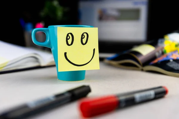 Smiling face drawn on sticky note on a cup of coffee at the office desk. Every morning wake up with the smile