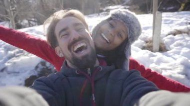 Çok ırklı bir çift, karlı dağlarda tatildeyken selfie çeker. Kış tatili genç adam ve kadın ormanda doğanın tadını çıkarıyorlar. Yüksek kalite 4k görüntü