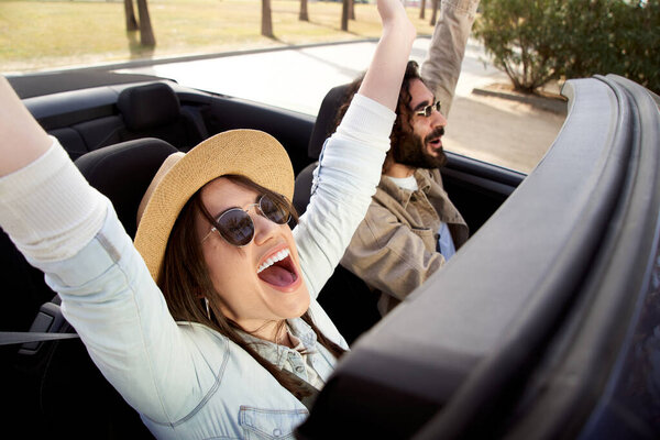 Happy couple friends enjoying dancing in the car. Sitting in front seat and having fun on a road trip. Man and woman driving and dancing. Concept of youth, relationship, holidays and vacation.