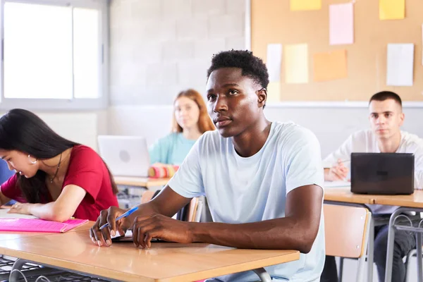 African American student attending teacher lesson at classroom. Black guy in class study with a multiracial high school college. Young boys and girls sitting at desk. Education concept. High quality