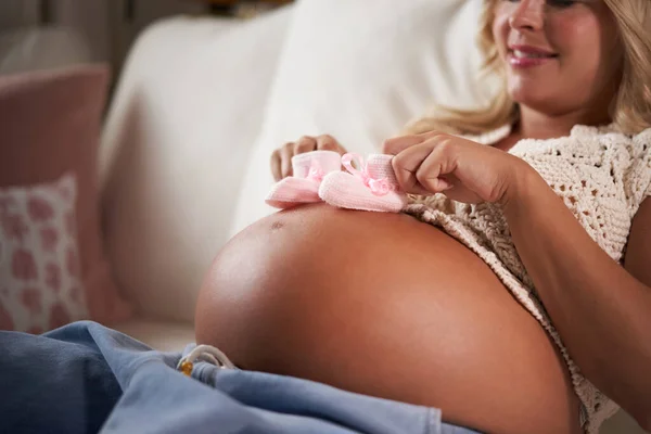 Close-up of an unrecognizable pregnant mom touching her belly and newborn shoes sitting on the couch at home. Woman in the last stage of pregnancy waiting for the birth of her baby.