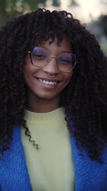 Vertical Close up portrait of beautiful smiling young African woman with curly hair looking at camera with positive expression. Happy latin girl wearing eyeglasses outdoors. Cheerful real people.