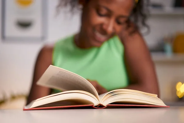 Young latin woman smiling indoors at home reading a book. Pretty African American girl sitting in a desk. People passionate about literature. Close up. Selective Focus on the book.