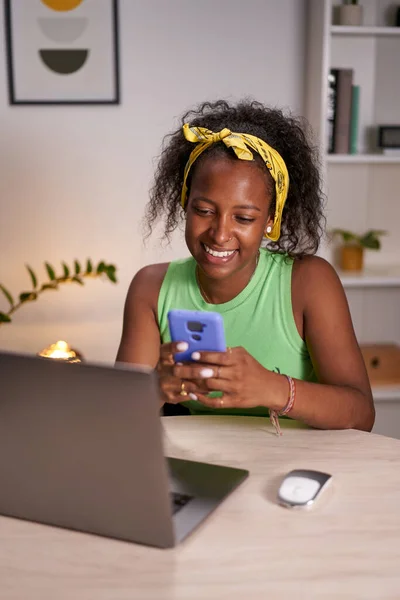 Vertical photo of a Young African woman using phone at home office. Happy girl typing on the mobile cell and telecommuting. Cheerful sitting at desk. Positive thinking person concept.