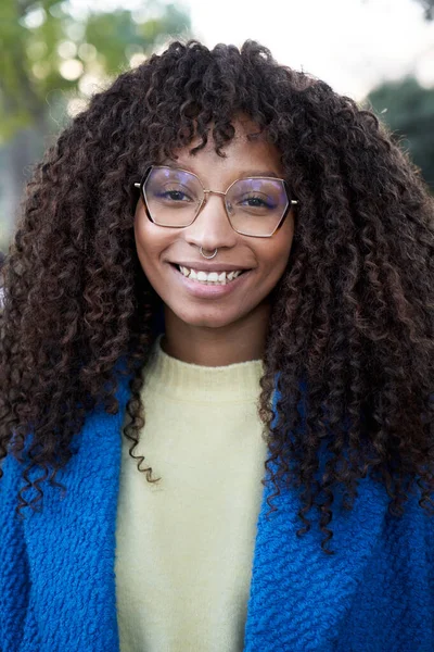 Vertical Close up portrait of beautiful smiling young African woman with curly hair looking at camera with positive expression. Happy latin girl wearing eyeglasses outdoors. Cheerful real people.