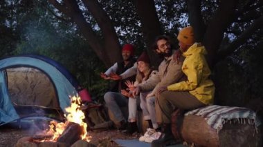 Group of people spending free time and sitting during camping together on winter day. Cheerful young travelers talking and warming each others hands at the bonfire. Lifestyle moment at nature.