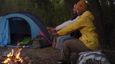 African people spending free time and sitting during camping together on winter day. Young couple of travelers talking and warming each others hands at the bonfire. Lifestyle moment at nature.