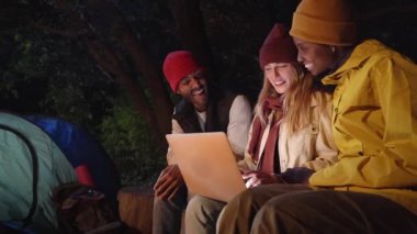 Friends using computer during camping together on winter night. Happy young people making a video call. Camping tent for outdoor holiday and vacation. Lifestyle concept.