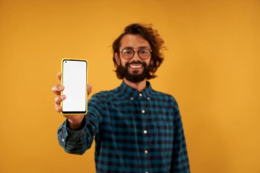 Young bearded man in glasses shows on blank smartphone screen isolated on yellow background. Guy in blue plaid shirt posing on studio portrait. Copy space. Male holding mobile phone.