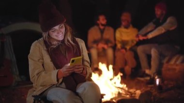 Young blonde girl typing on your mobile on the background of a campsite at night in outdoors. Millennial woman full of joy chatting with someone on mobile phone sitting in front of the bonfire. 