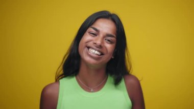 Portrait of Young Indian Woman Looking at Camera and Smiling in Color Studio Shot. Happy adult girl isolated on yellow background having fun Closeup. Indian person with beautiful skin tilts head.