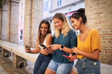 Dışarıda cep telefonu kullanan üç mutlu kadın. Bir grup gülümseyen bayan arkadaş akıllı telefondan sosyal medyayı izliyor. Yüksek kalite fotoğraf