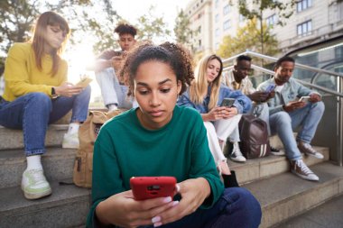 Focused girl looks and types on her smart phone sitting on city street with friends. Serious young millennial youth in casual clothes technology addicted outdoor background. Sunny day with classmates