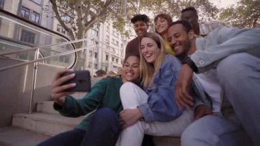 Group of young people taking a selfie or video with their phones sitting on the stairs. Smiling multiracial friends posing for a photo outside the University campus. Students in their free time.